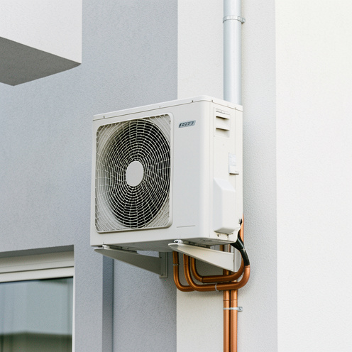 Unité extérieure compacte de climatisation installée contre le mur d’un appartement moderne, avec grille de ventilation et conduites isolées visibles.